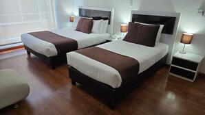 Quarto de hotel com duas camas, piso de madeira, mesas de cabeceira com luminárias e uma janela com persianas.
