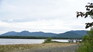Vista a la orilla de un lago con una mesa de picnic y bancos, rodeado de montañas y un área boscosa.