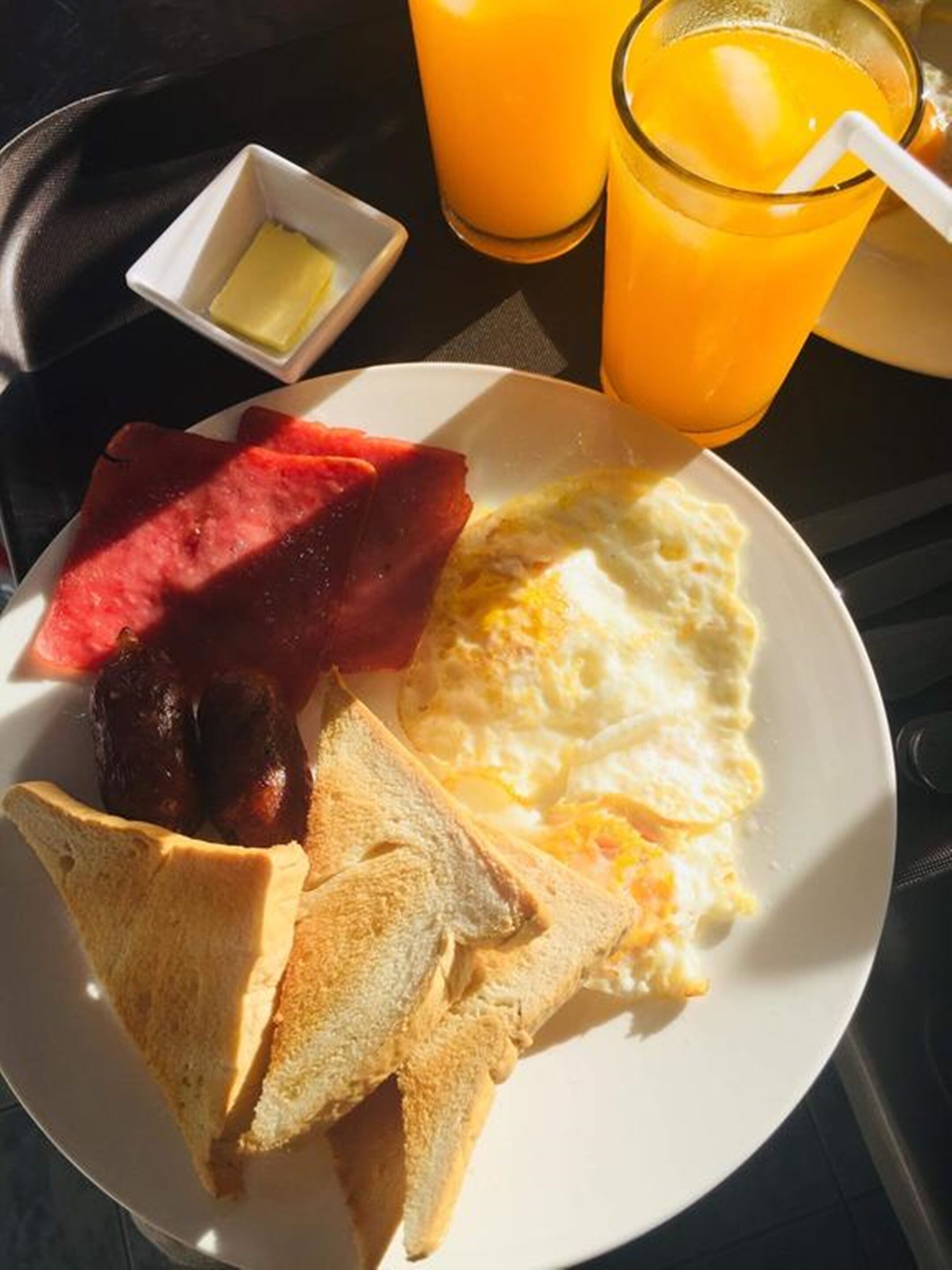 Een ontbijt bord met toast, eieren, ham en een stukje watermeloen. Ook staan er twee glazen jus d’orange op tafel.