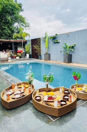 A poolside area with wicker furniture containing food and drinks, surrounded by potted plants and a building in the background. - Villa Marii (Dipolog)