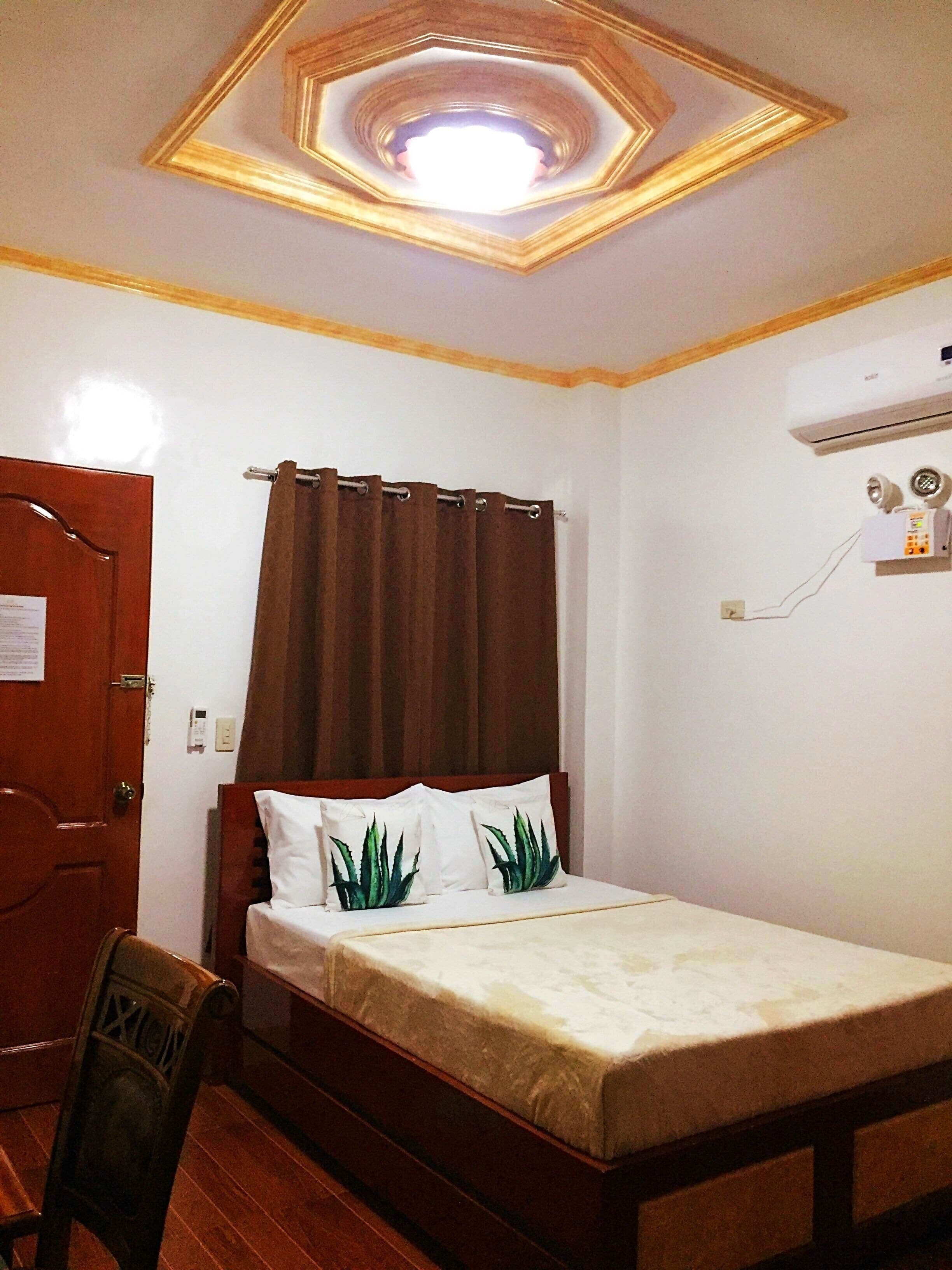 Una habitación de hotel con cama, una silla de madera, una puerta, una cortina, un equipo de aire acondicionado y un aplique en el techo.
