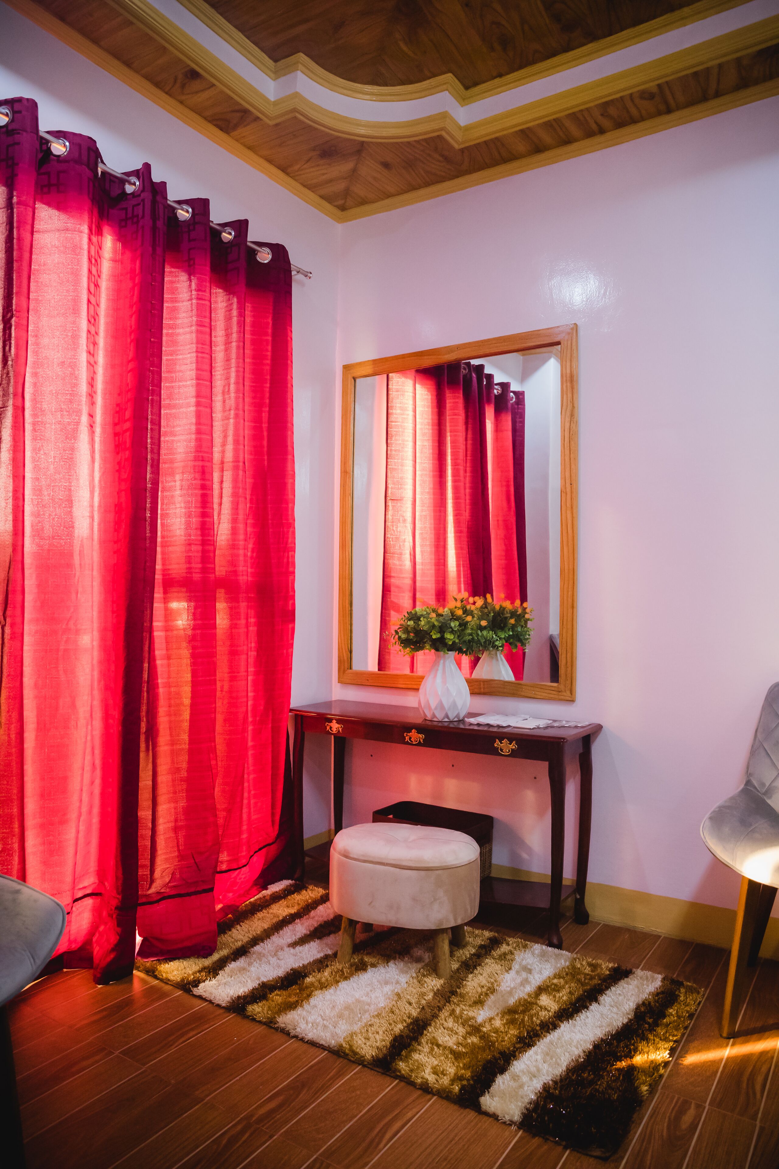 A room with a wooden ceiling, a mirror, a vanity with a vase of flowers, a striped rug, and a chair.