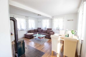 A modern living room with a brown sofa, glass coffee table, and a white armchair. A black wood-burning stove is visible in the corner.