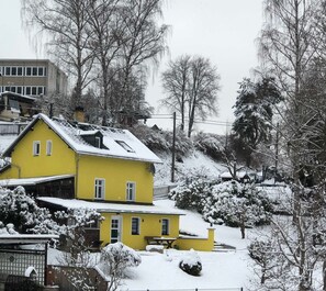 A yellow house with a sloped roof, surrounded by snow-covered trees and bushes.