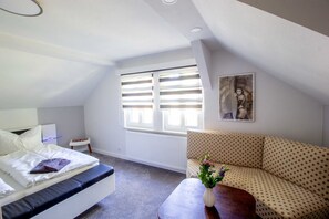 Un dormitorio con una cama, un sofá, una mesita con un florero y una ventana con persianas.
