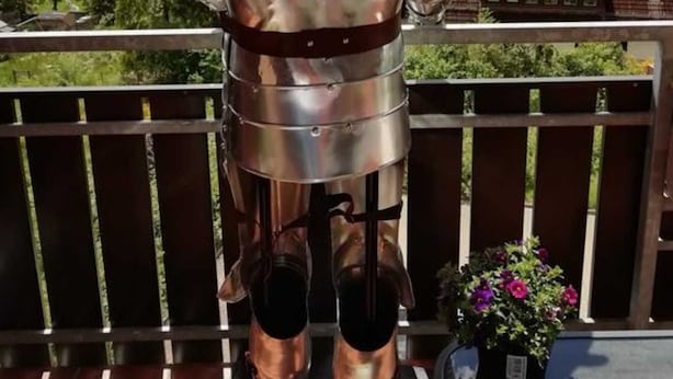 Une statue d’un chevalier en armure complète, placée sur un balcon d’où l’on peut admirer des maisons et des arbres.