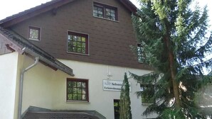 Il s’agit d’un bâtiment à deux étages, sur lequel on peut voir une enseigne indiquant qu’il s’agit d’une maison d’hôtes. Une clôture en bois se trouve devant, et un grand arbre à feuilles persistantes se dresse sur la droite.