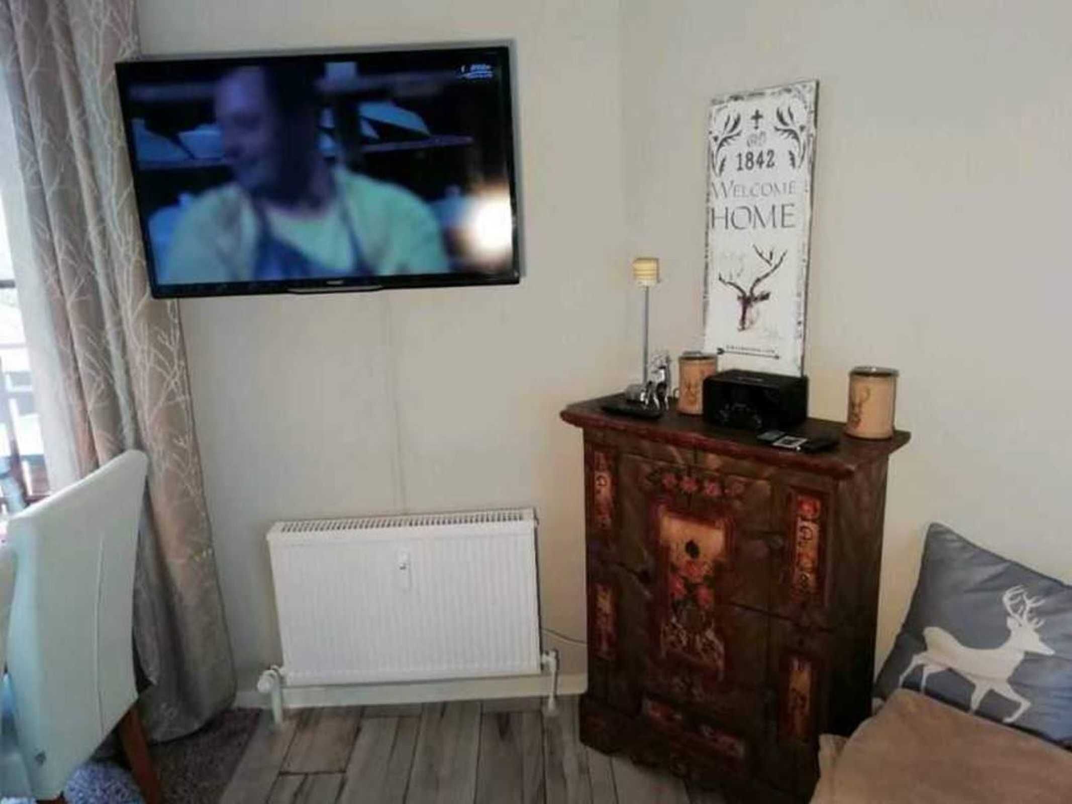 Una sala de estar con un televisor montado en la pared, un radiador, un mueble de madera y un letrero decorativo.