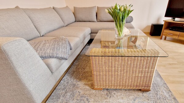 A modern living room with a grey sofa, a glass coffee table, and a wicker side table.