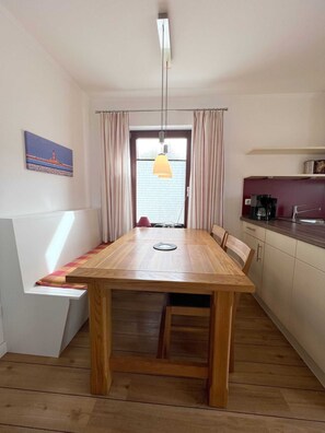 A dining area with a wooden table and chairs, a pendant light, a painting on the wall, and a kitchen area with cabinets and a countertop.