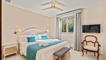 Un dormitorio con cama, mesitas de noche, un banco, una silla, televisor y ventana con cortinas.