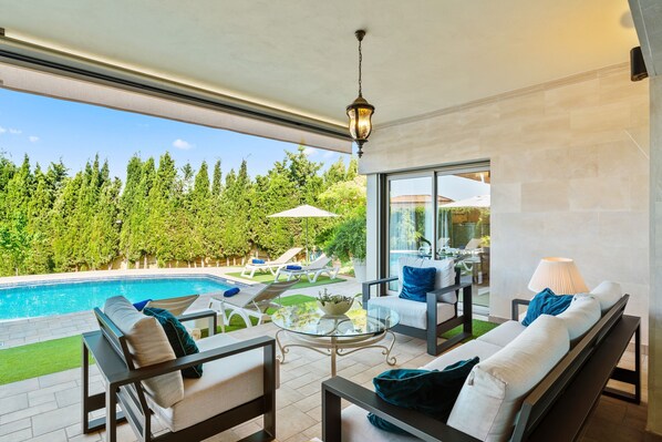A modern outdoor living area with a pool, lounge chairs, a glass table, and a view of a forested area.
