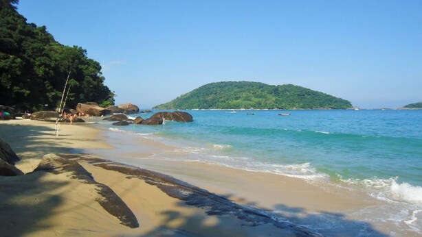 Uma praia com águas azul-claras, areal arenoso e uma ilha coberta por floresta ao fundo.
