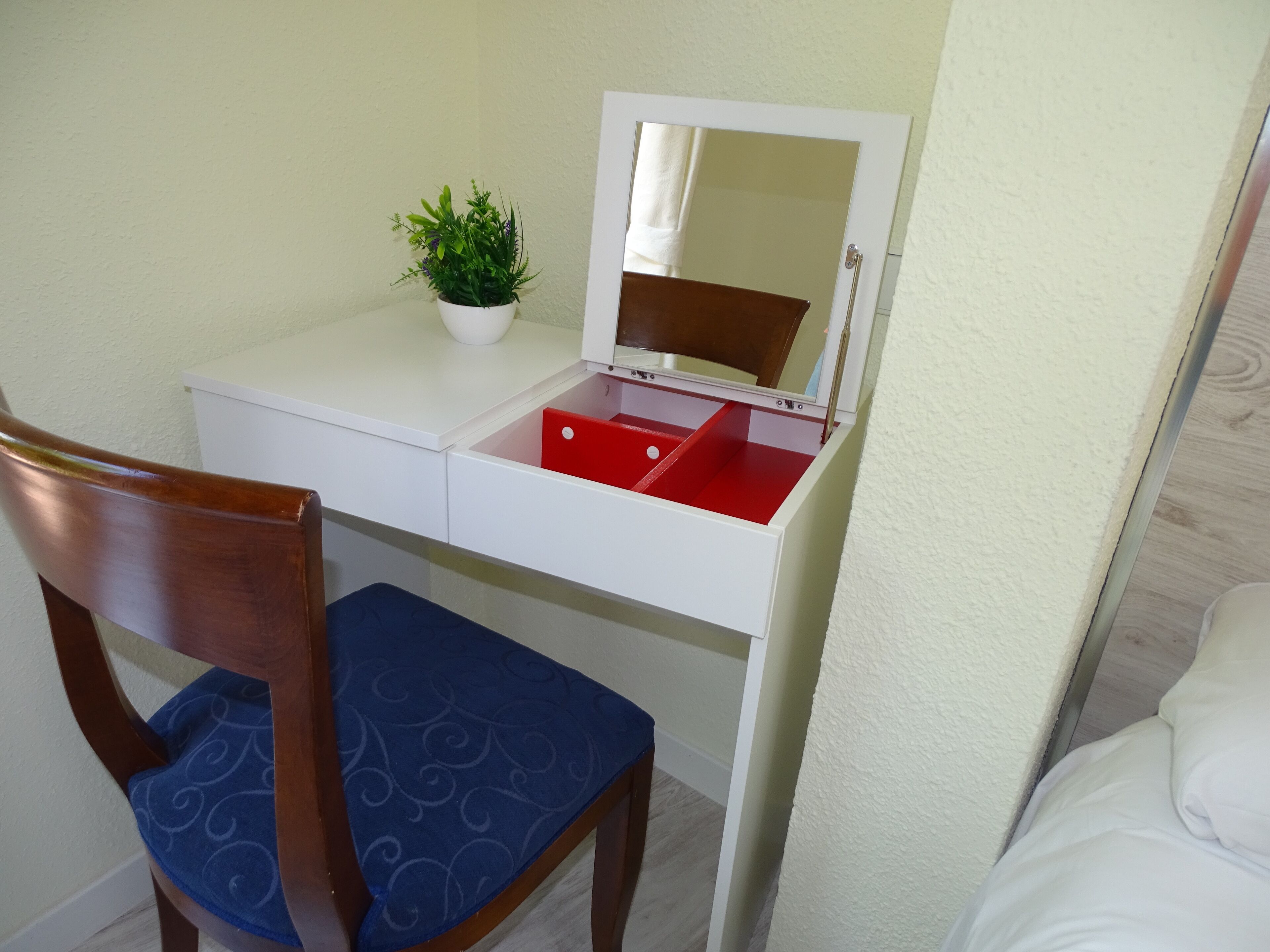 Una habitación pequeña con un escritorio blanco, un cajón rojo, una silla de madera con cojín azul y una pequeña planta en maceta.
