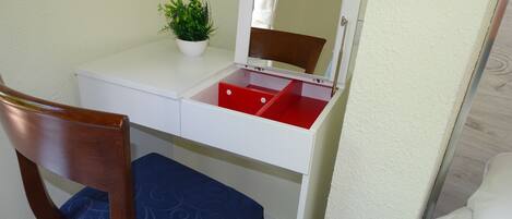 Una habitación pequeña con un escritorio blanco, un cajón rojo, una silla de madera con cojín azul y una pequeña planta en maceta.