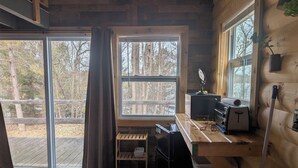 A cabin interior with a wooden desk, a black refrigerator, a window with curtains, and a view of trees outside.