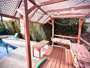 A wooden deck with a pergola, a pool, and a lounge area with cushions.