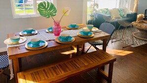 Een eethoek met een houten tafel voor vier personen, omringd door stoelen. Een groot raam met zonwering is zichtbaar, en er staat een potplant op de vensterbank.