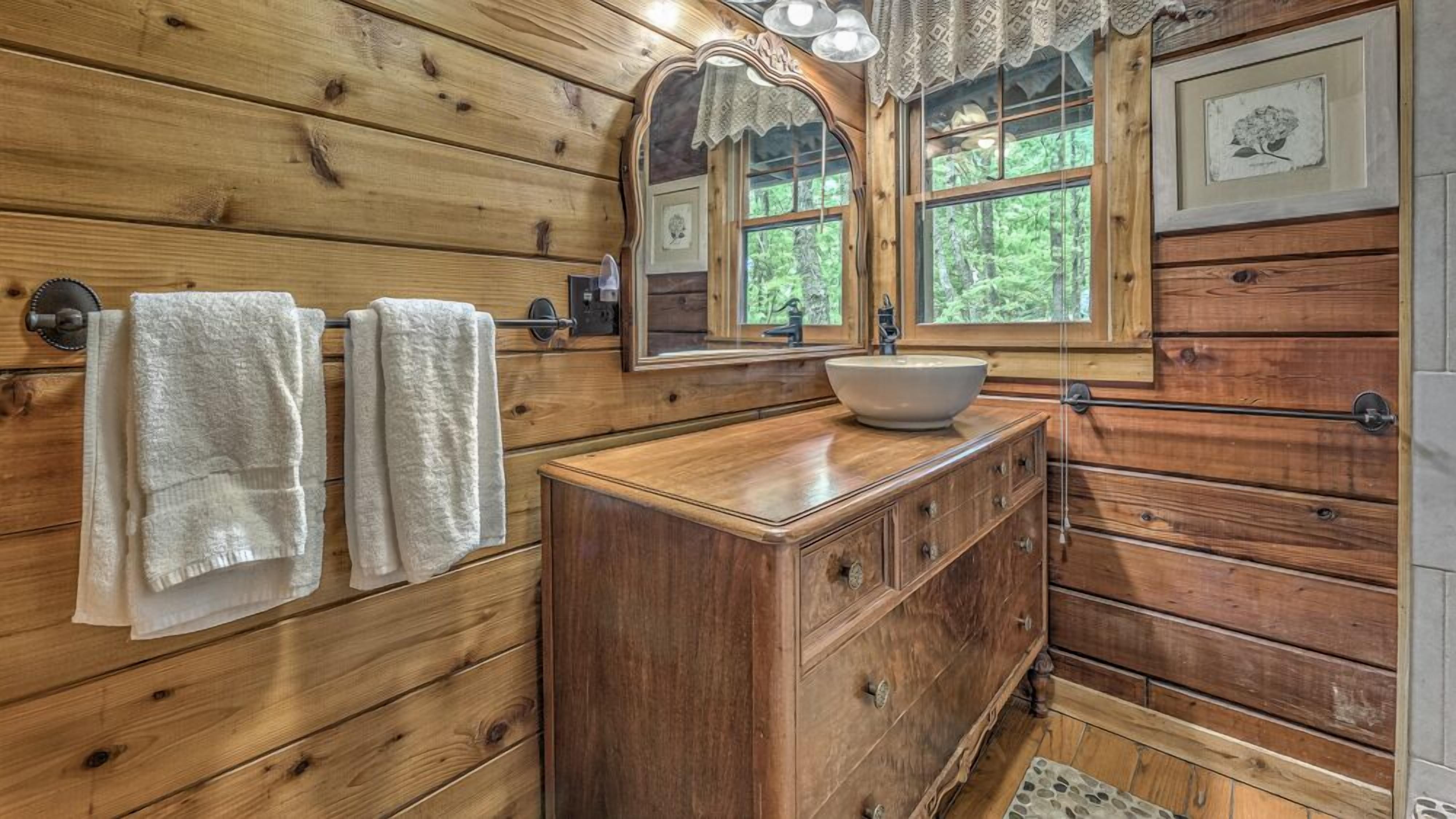 Baño de cabaña de madera con tocador de madera, un espejo grande y una ventana con vistas al bosque.