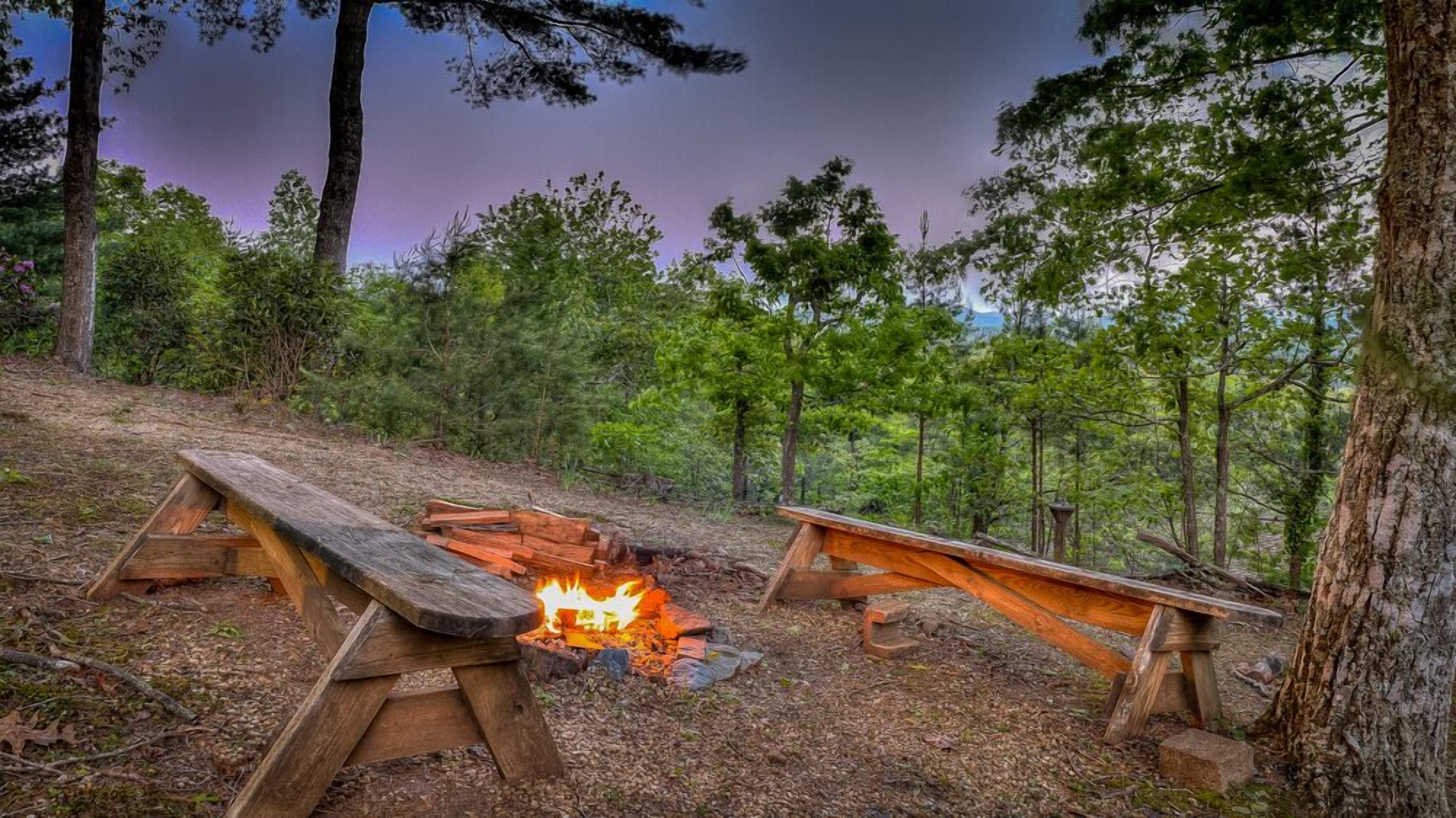 A campsite with two wooden picnic tables, one with a fire in the center.