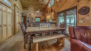Interior de una cabaña de madera con una mesa de comedor, sillas, un banco, una zona de cocina y un ventanal grande.