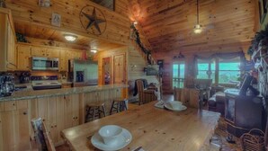 A wooden cabin interior with a kitchen, dining area, and a large window overlooking a scenic view. - Heavens Whisper- Lake and Mountain Views | Outdoor Fire Pit | Aska Adventure Area (Blue Ridge)