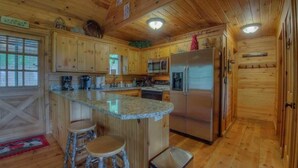 A wooden cabin kitchen with a central island, bar stools, a refrigerator, and a microwave. - Heavens Whisper- Lake and Mountain Views | Outdoor Fire Pit | Aska Adventure Area (Blue Ridge)