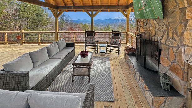 A covered outdoor seating area with a stone fireplace, a television mounted on the wall, and a view of the mountains in the background. - Heavens Whisper- Lake and Mountain Views | Outdoor Fire Pit | Aska Adventure Area (Blue Ridge)