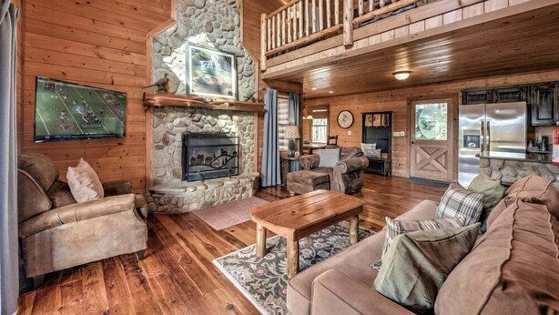A cozy cabin interior with a stone fireplace, wooden furniture, and a kitchen area. - Daze Off- Spectacular Views | Large Hot Tub | Outdoor Fire Pit (Morganton)