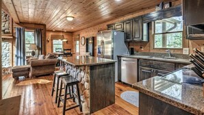 A rustic cabin interior with a stone fireplace, wooden floors, a dining area, and a kitchen with granite countertops. - Daze Off- Spectacular Views | Large Hot Tub | Outdoor Fire Pit (Morganton)