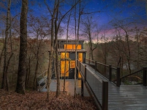 A cabin with a wooden deck and a staircase leading to a glass door, surrounded by trees.