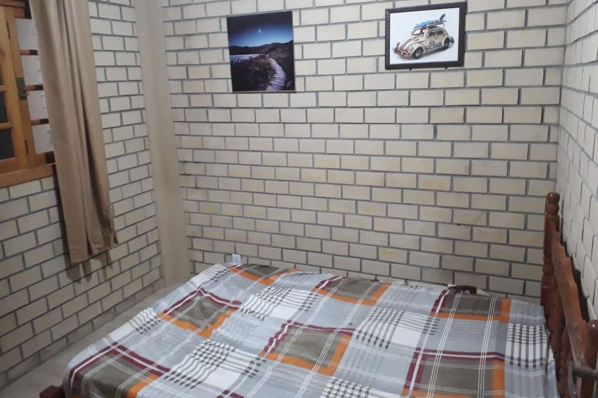 A small, narrow room with a brick-patterned wall, a bed with a plaid bedspread, and two framed pictures.