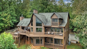 A two-story log cabin with a balcony and a deck, surrounded by dense forest.