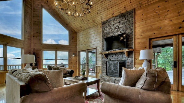 A spacious living room with a stone fireplace, large windows, and wooden walls.