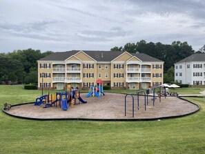 Children can play on the playground's slides and climbing structures surrounded by a lush green area.
