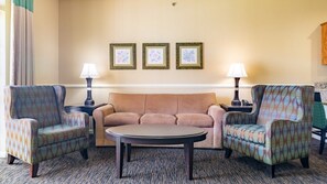 A hotel room with a beige sofa, two patterned armchairs, a round coffee table, and three framed pictures on the wall.