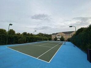 An outdoor tennis court with a blue surface, surrounded by a fence and trees, with a building in the background.
