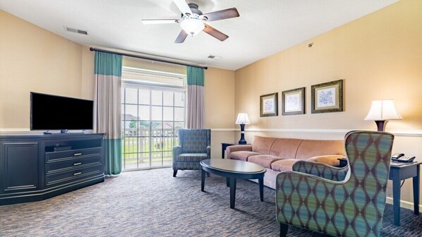 Ein Hotelzimmer mit Fernseher, einer Couch, einem gemusterten Sessel, einem Couchtisch und einem Deckenventilator.