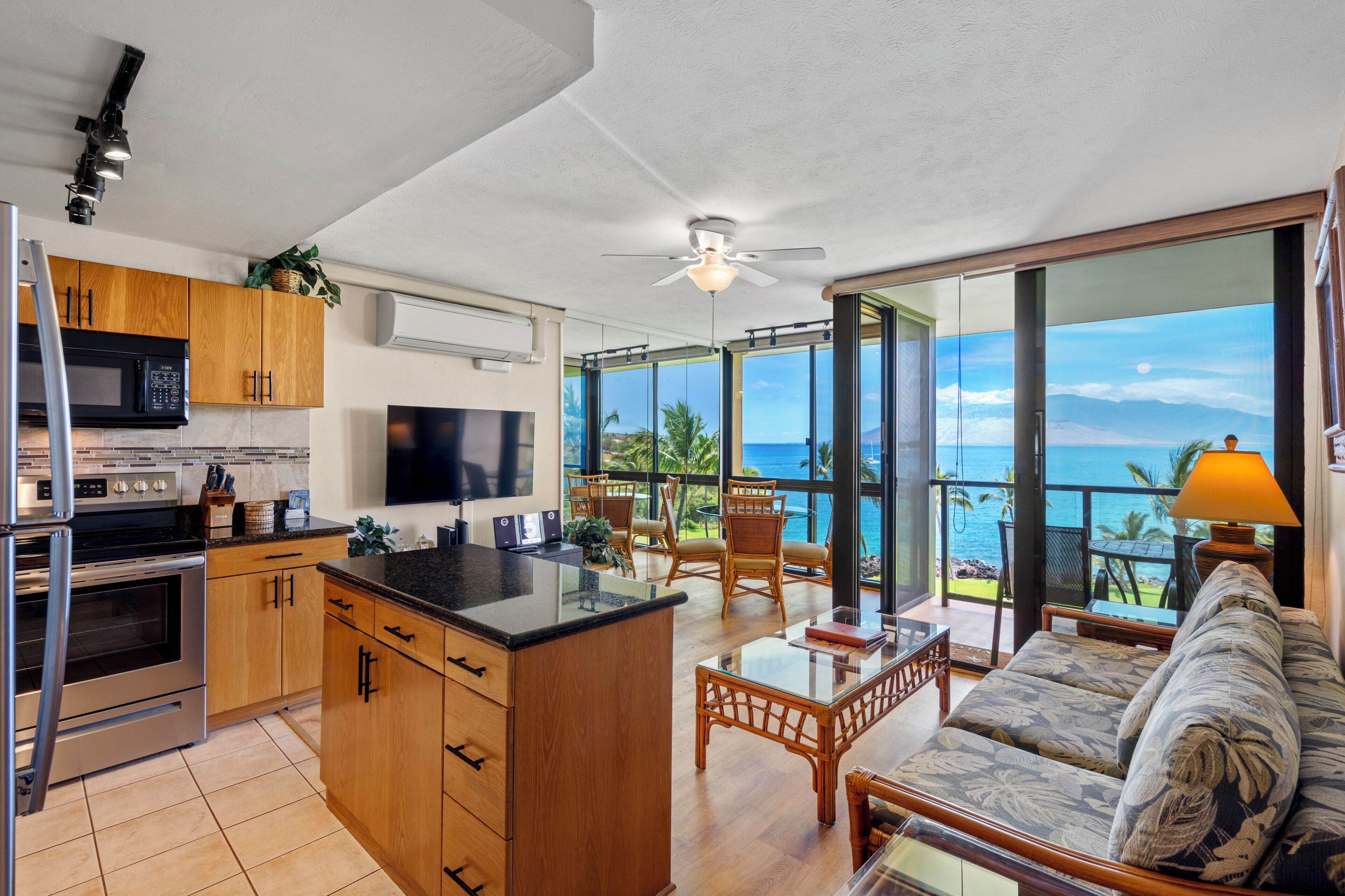A modern kitchen with wooden cabinets, a large island, and a flat-screen TV. There is a dining area with a glass table and chairs, and a sofa with a view of the ocean.