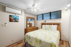 Ein Schlafzimmer mit Bett, Nachttisch, Spiegel, Deckenventilator und Fenster mit Jalousien.