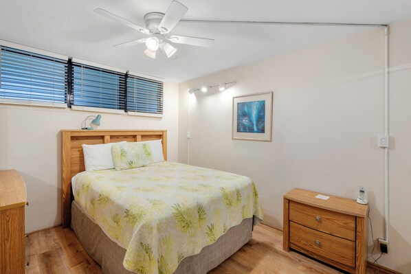 Ein Schlafzimmer mit einem Bett, einem Nachttisch, einem Deckenventilator, einem Fenster mit Jalousien und einer Wandleuchte.