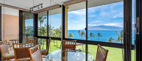 A room with a glass table surrounded by wicker chairs, large windows with a view of the ocean and palm trees, and a wooden ceiling.