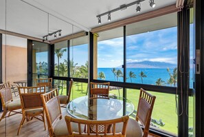 A room with a glass table surrounded by wicker chairs, large windows with a view of the ocean and palm trees, and a wooden ceiling.