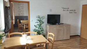 Une pièce comprenant une table à manger, des chaises, un téléviseur posé sur un meuble en bois, une plante en pot et un mur sur lequel figure une liste de noms de villes.