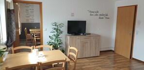 Een kamer met een eettafel, stoelen, een televisie op een houten kast, een potplant en een muur met een lijst van steden.