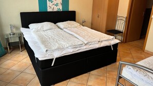 Un lit avec un cadre noir, un matelas blanc et une couette blanche. La chambre contient une petite table de chevet avec une lampe, une chaise et une armoire.