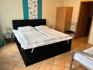 Een bed met een zwart frame, een witte matras en een witte dekbedovertrek. In de kamer staat een klein nachtkastje met een lamp, een stoel en een kledingkast.