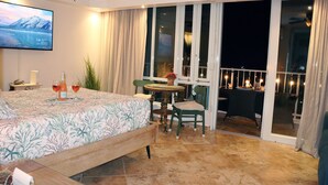 Habitación de hotel con cama, televisor, balcón con mesa y sillas, y vistas al cielo nocturno.