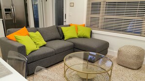 Un salon moderne avec un canapé d’angle gris, une table basse en verre et un pouf à motifs.