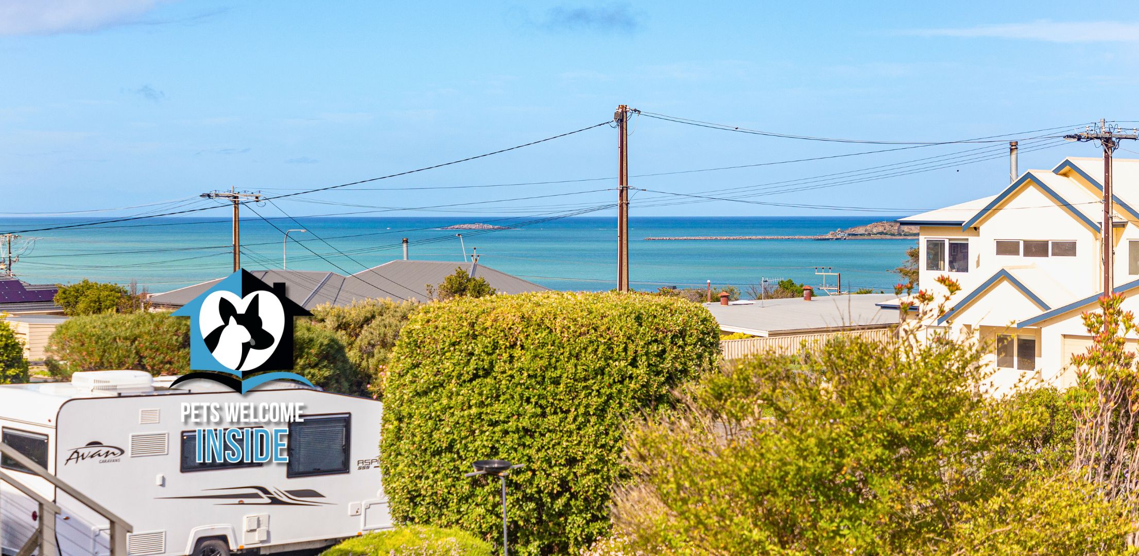 A caravan with a 'PETS WELCOME INSIDE' sign, surrounded by greenery and overlooking a coastal area with houses and a clear sky.